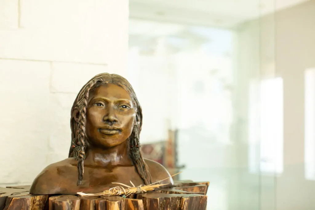 A bronze bust of a woman with braided long hair on a wooden stand indoors, background blurred with glass and white walls.