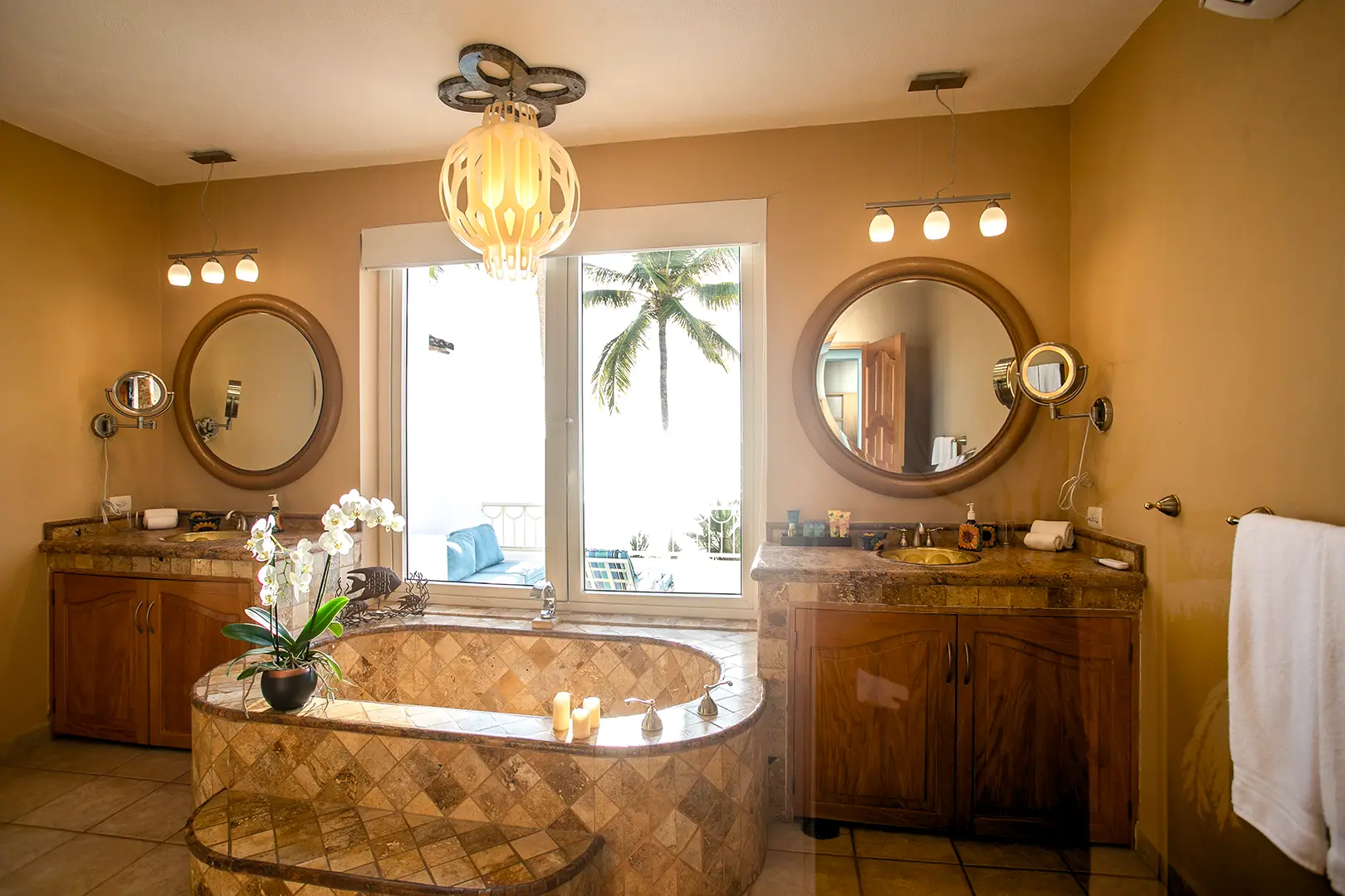 Bathroom with central oval tub, two wooden vanities, round mirrors, orchid, candles, and a window view of palm trees.