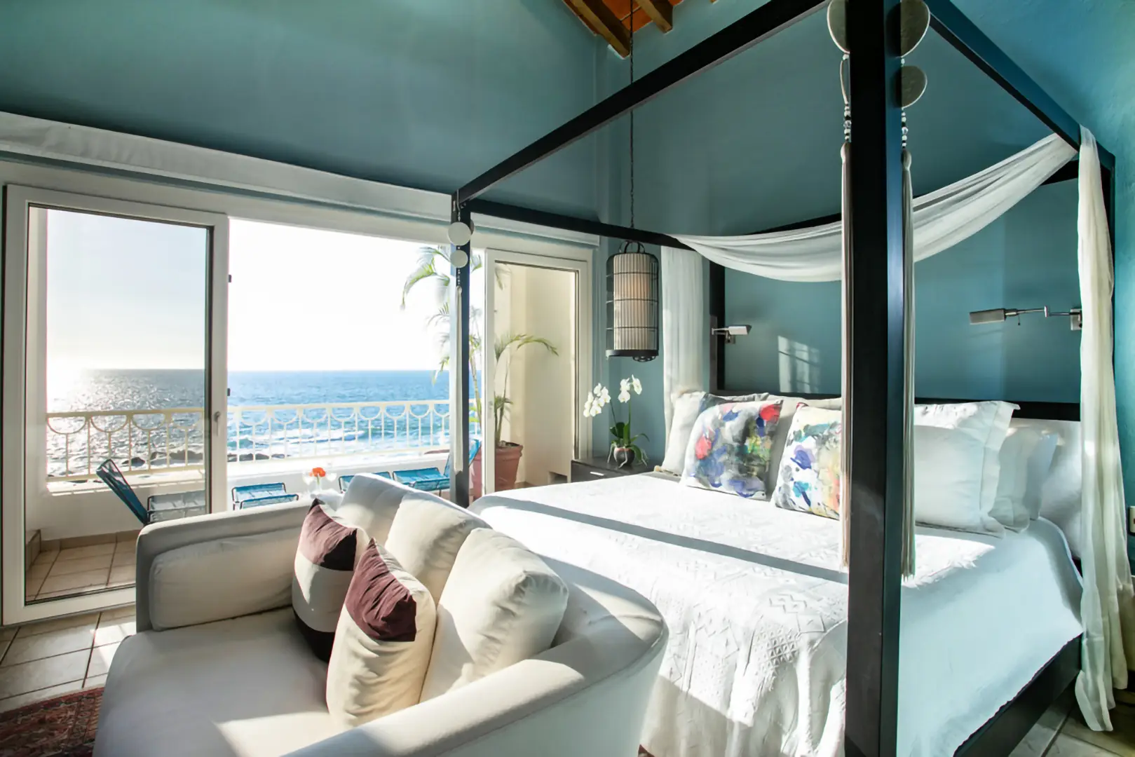 Seaside bedroom with four-poster bed, white sofa, blue walls, and balcony facing ocean and lounge chairs.