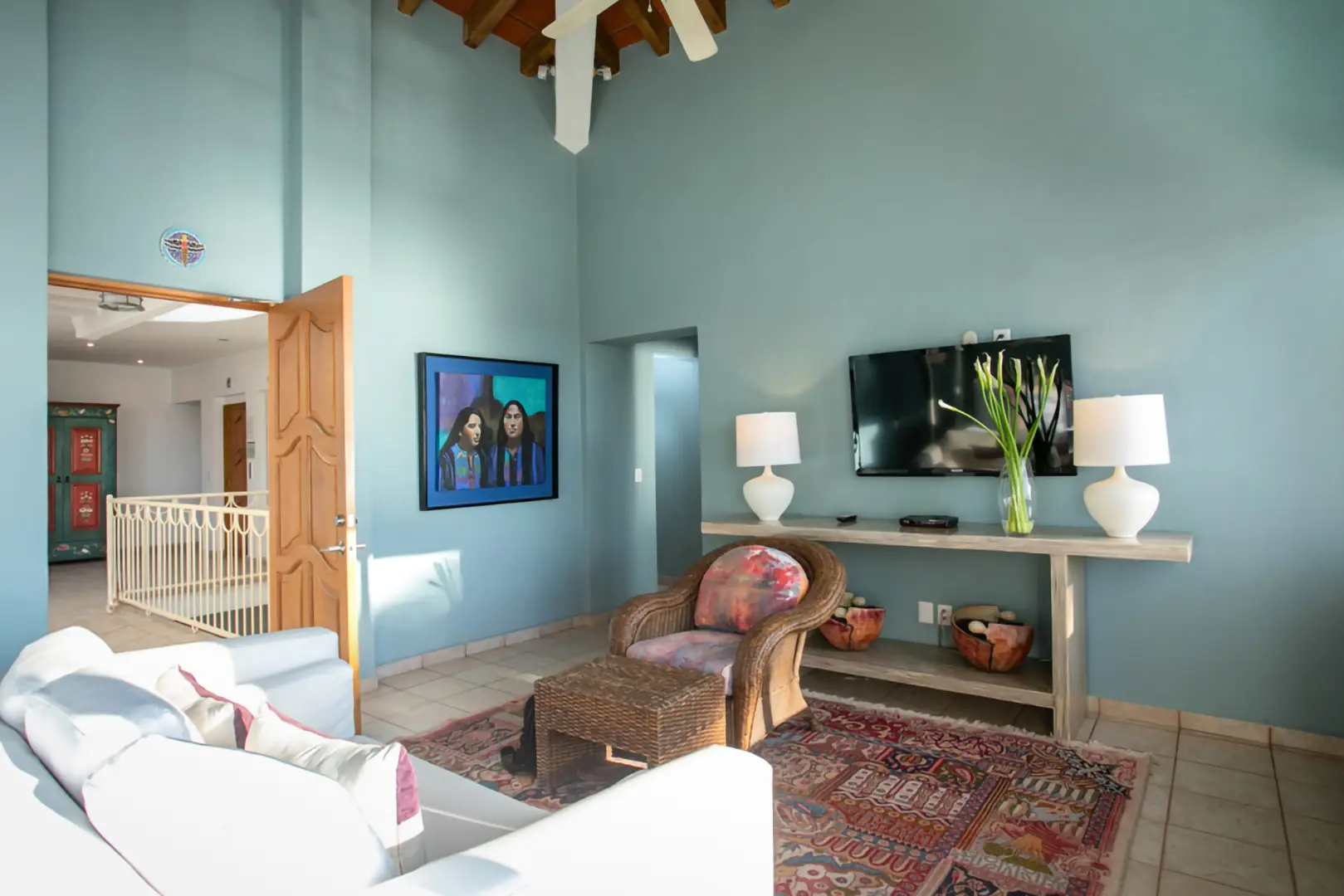 Bright living room with blue walls, white sofa, wicker chair, colorful rug, TV, two lamps, console table, and artwork.