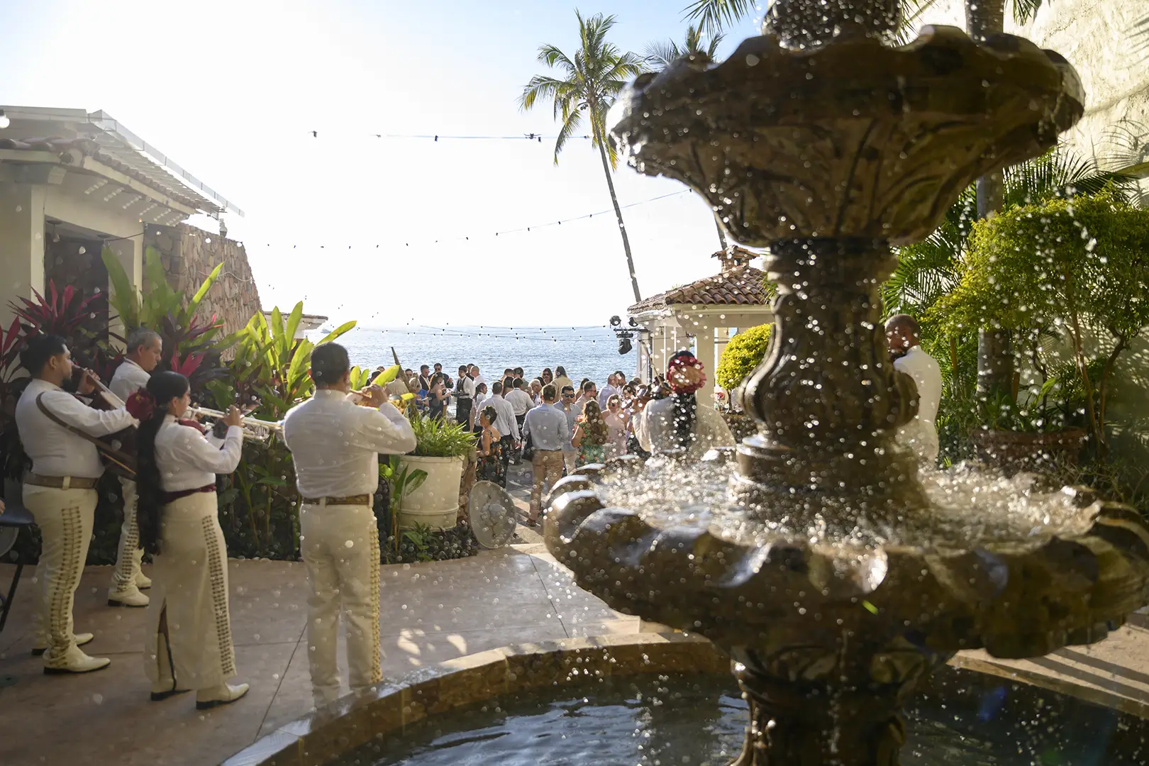 A mariachi band plays by a fountain at an outdoor wedding venue with guests, ocean view, palm trees, and a sunny sky.