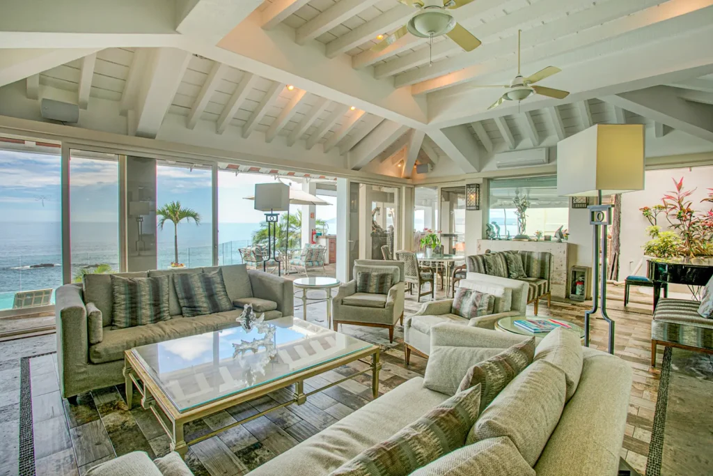 Sunlit living room with sofas, glass coffee tables, large ocean-view windows, plants, and ceiling fans.