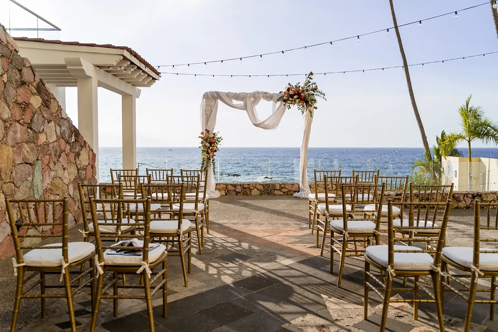 Outdoor wedding ceremony with gold chairs facing a white floral arch, string lights, and ocean view under sunny sky.