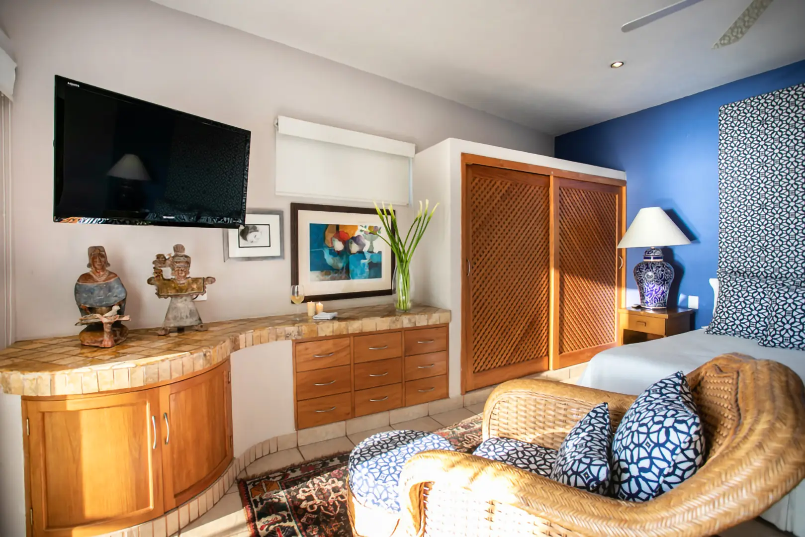Bedroom corner with TV above wooden desk, sculptures, framed art, vase with flowers, wicker chair, wardrobe, and lamp.
