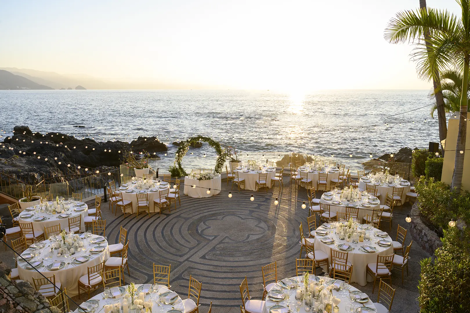 Outdoor wedding reception by the ocean at sunset with round tables, gold chairs, string lights, and a floral arch.