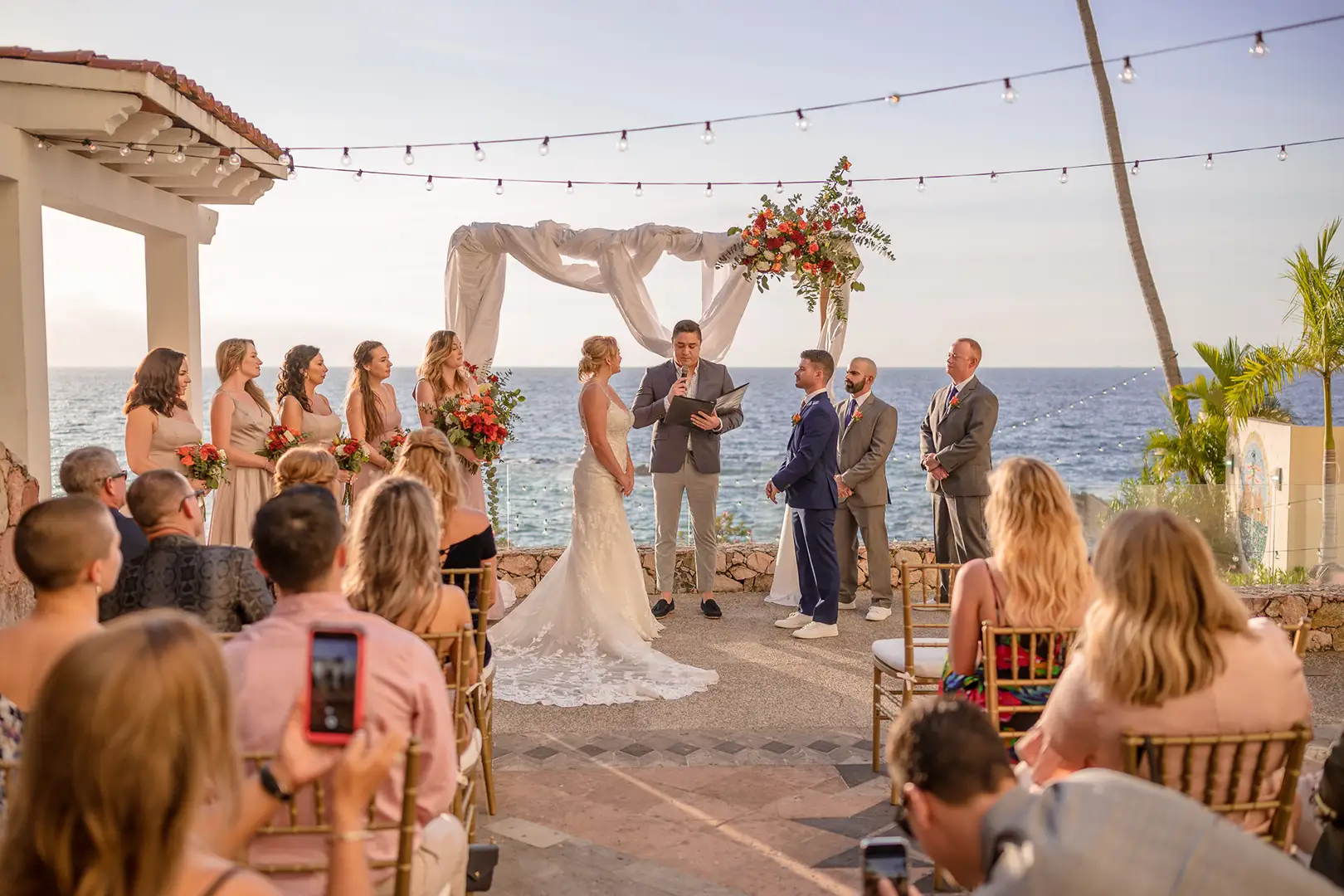 A bride and groom stand under a floral arch by the ocean, exchanging vows with attendants and seated guests nearby.