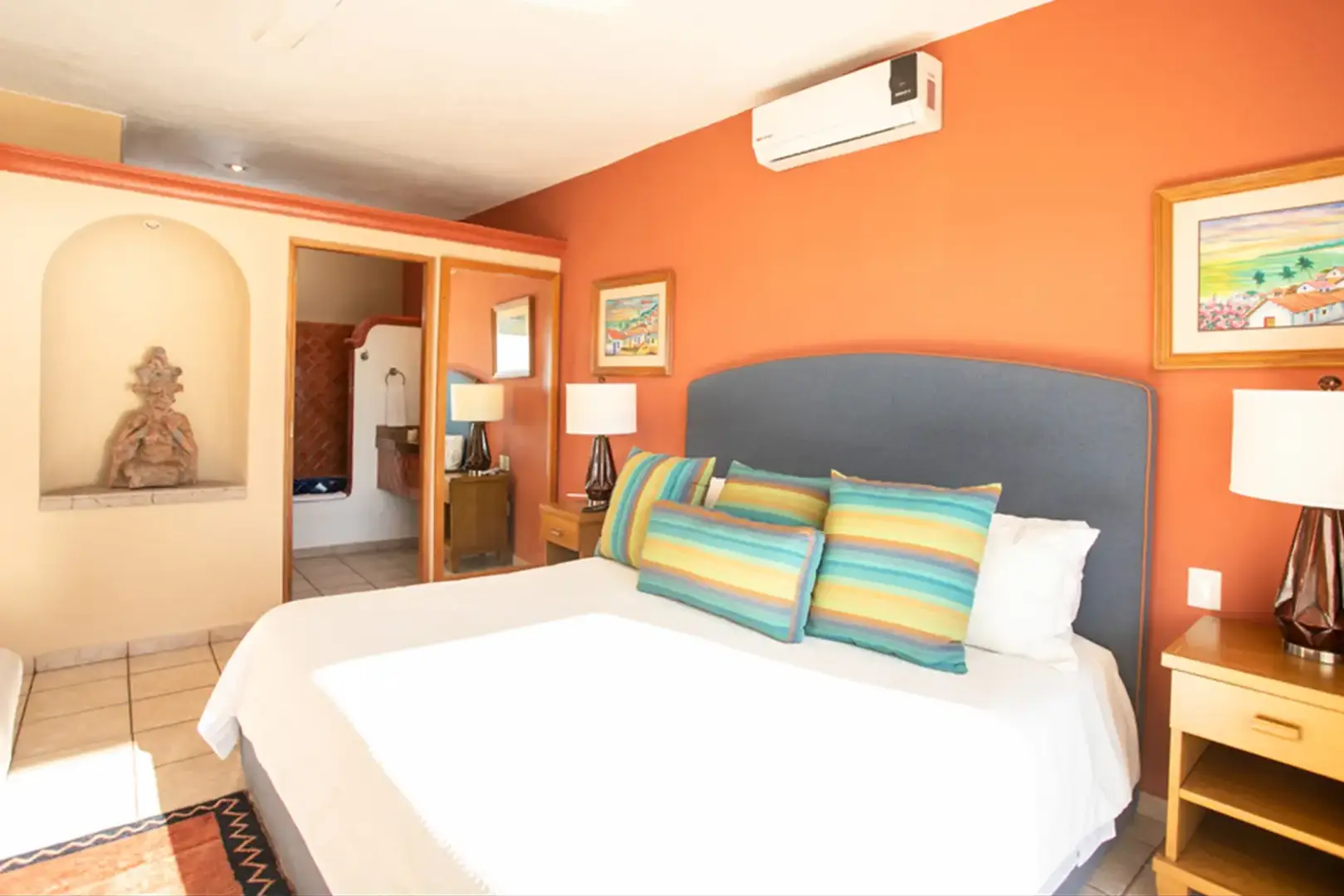 Bright bedroom with orange accent wall, large bed with striped pillows, bedside tables, lamps, art, AC, and open bathroom door.