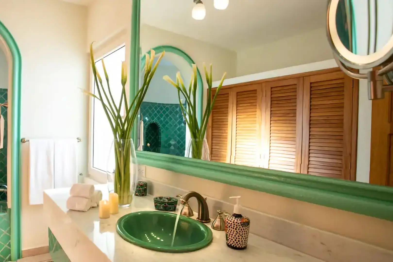 Bright bathroom with green sink, green-framed mirror, green tiles, white flowers, towels, candles, and closet doors.