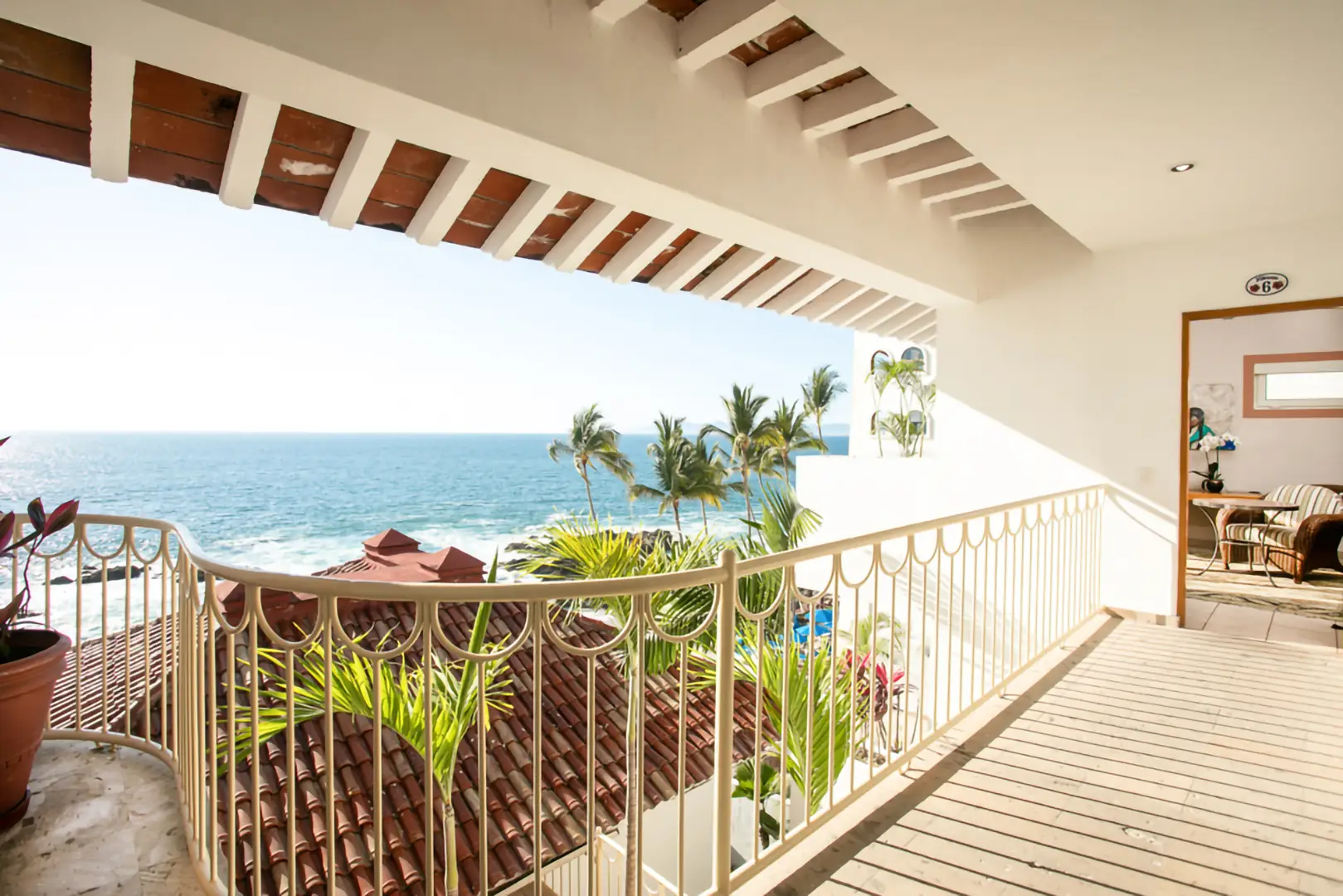 A sunlit balcony with a white railing overlooks red-tiled roofs, palm trees, and the ocean. Part is shaded by wood beams.