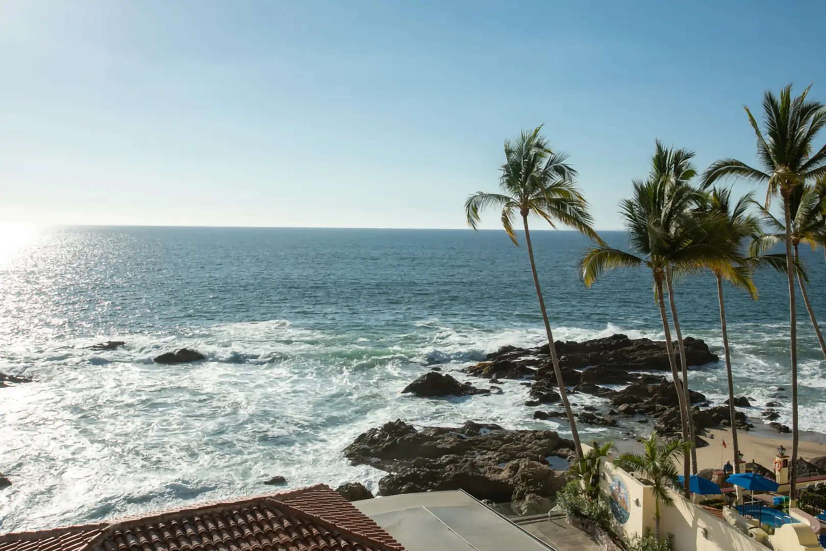 Ocean waves hitting rocky shore, two tall palm trees, and a red-tiled roof building under clear blue sky.