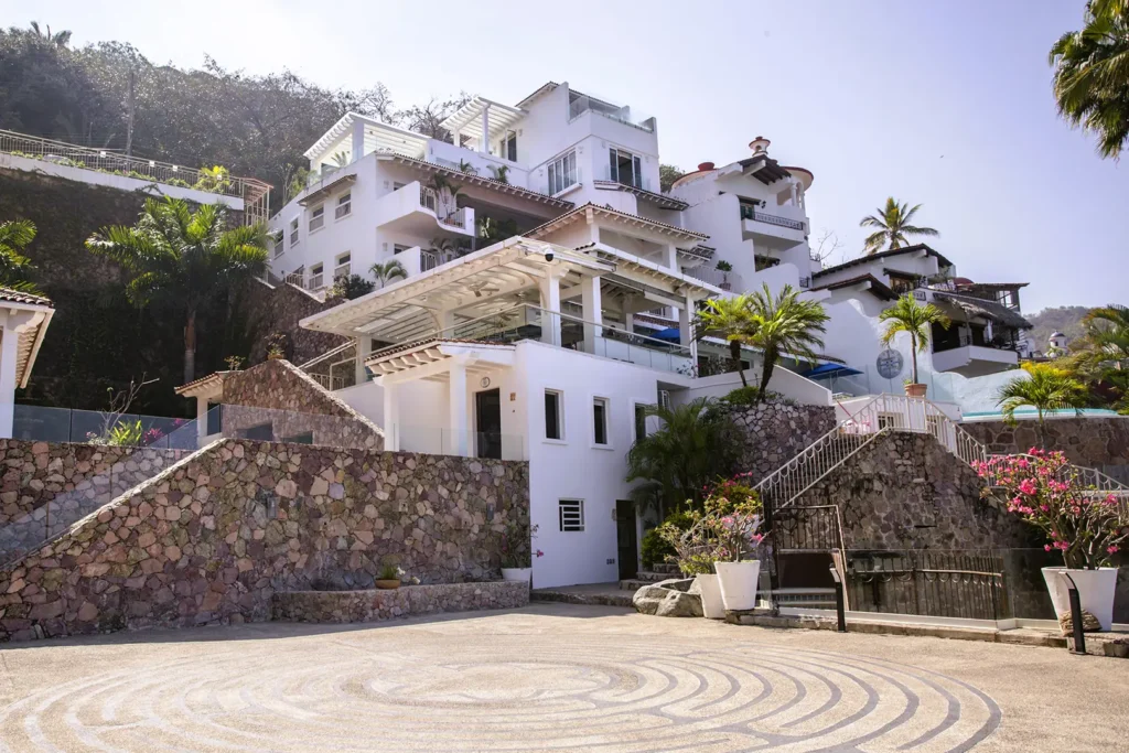 Multi-story villa with white balconies, palm trees, bougainvillea, stone wall, and circular courtyard in front.