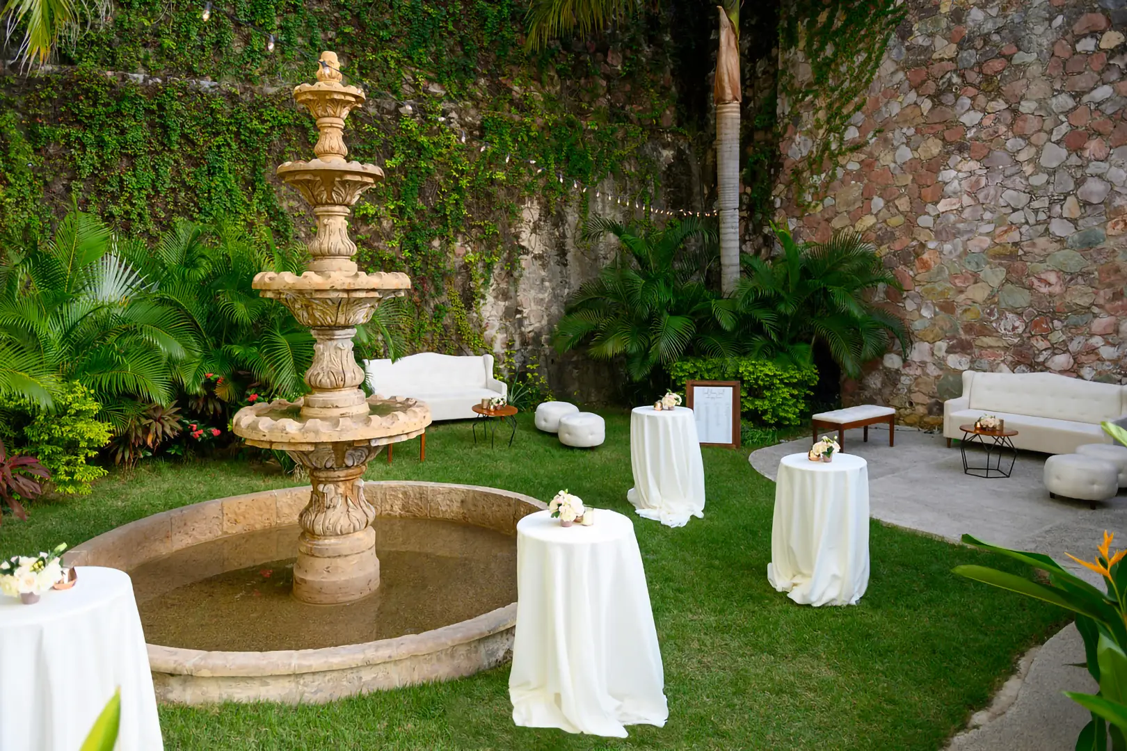 A stone fountain with small tables, white tablecloths, floral centerpieces, sofas, green plants, and a stone wall.