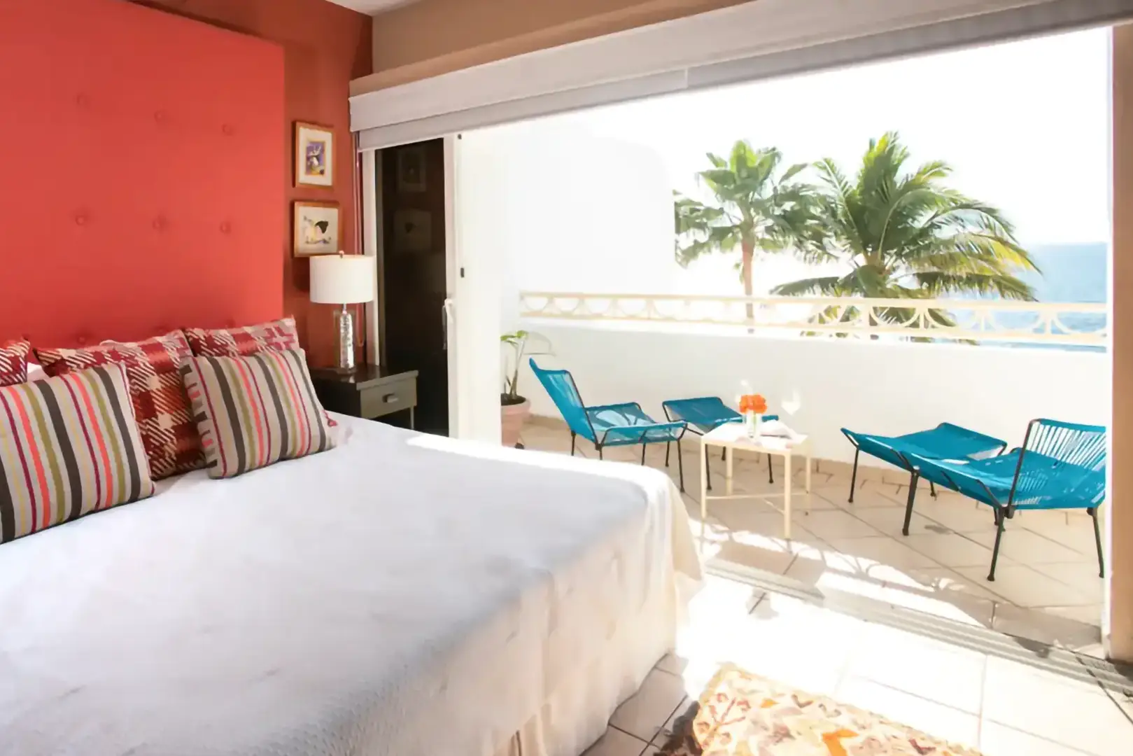 A bedroom with a large bed and colorful pillows opens to a balcony with blue chairs, table, palm trees, ocean view.