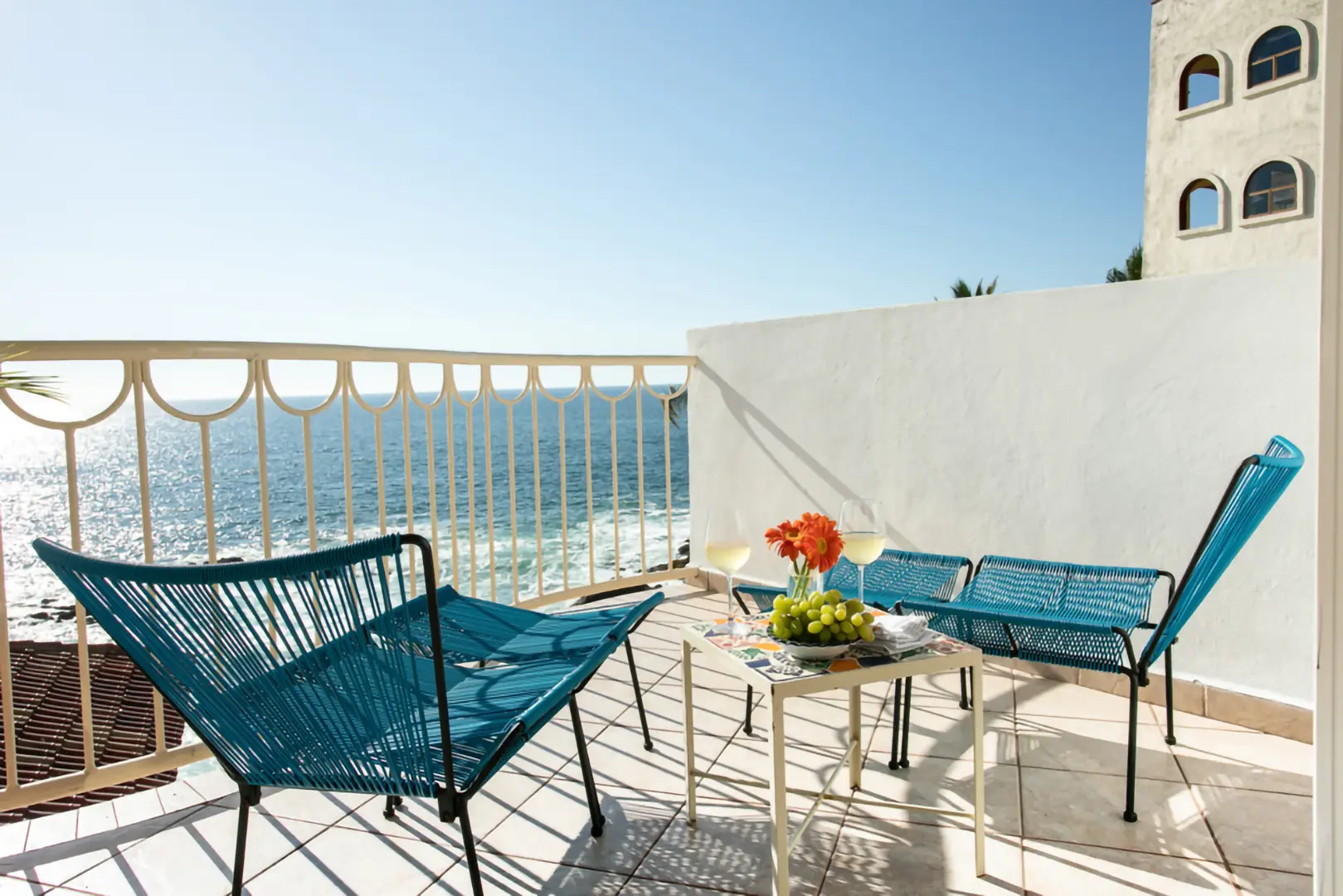 A patio with two blue chairs, a blue bench, a table with flowers, fruit, magazines, and wine overlooks the ocean.