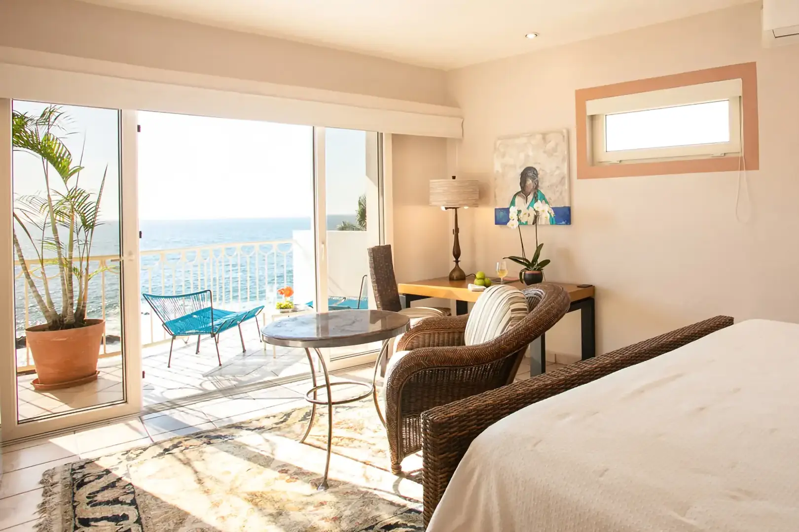 Bedroom with wicker chairs, round table, desk, bed, and sliding doors to balcony with blue chairs and ocean view.