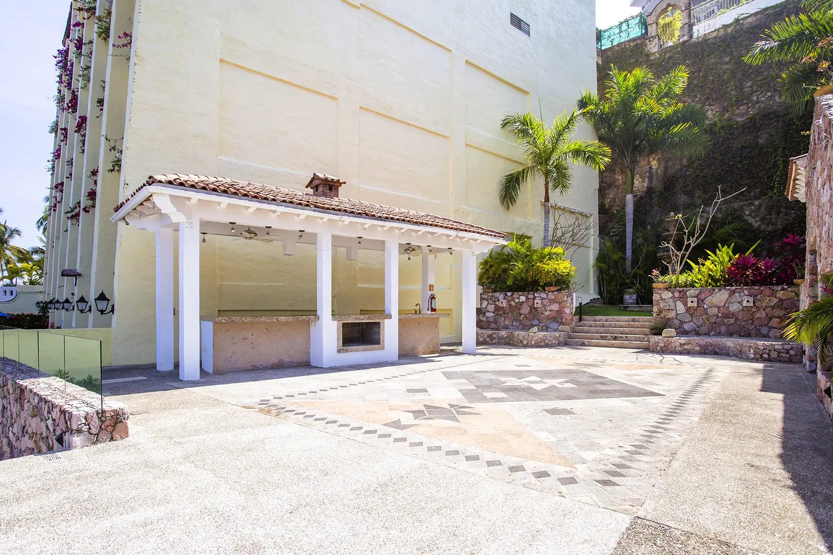 Outdoor patio with covered bar or grill, tiled floor, stone steps, tropical plants, and a light-colored building.