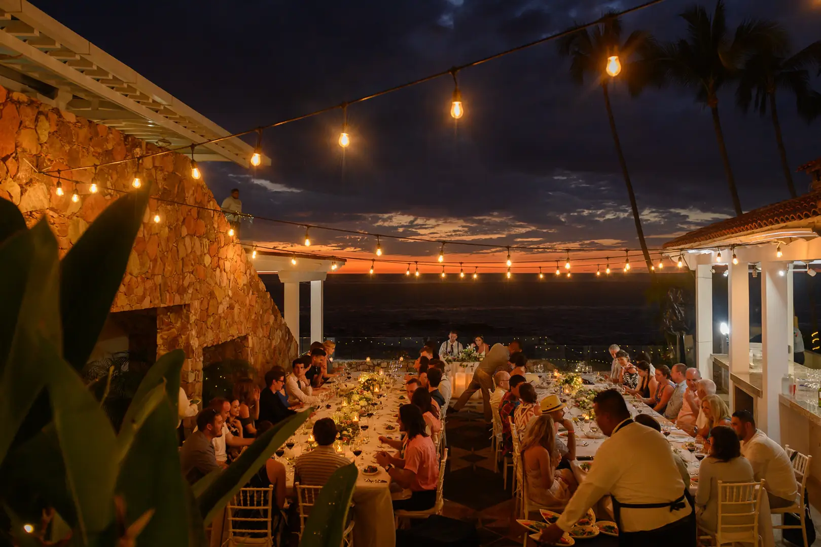 Guests seated at long tables outdoors at sunset, under string lights, with staff serving food. Stone walls and plants visible.