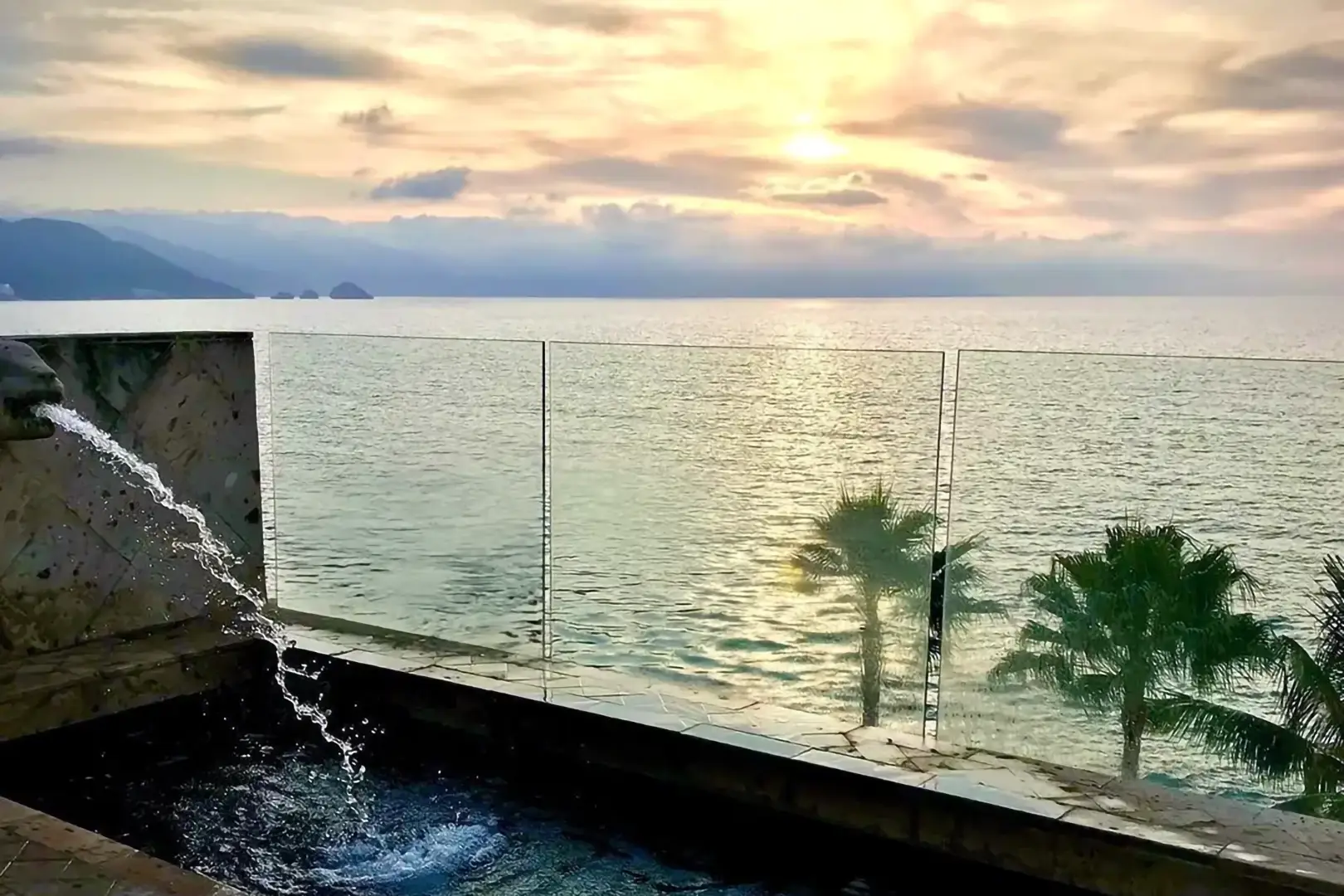Infinity pool with water streaming in, glass barrier, palm trees, calm ocean at sunset, mountains and clouds beyond.