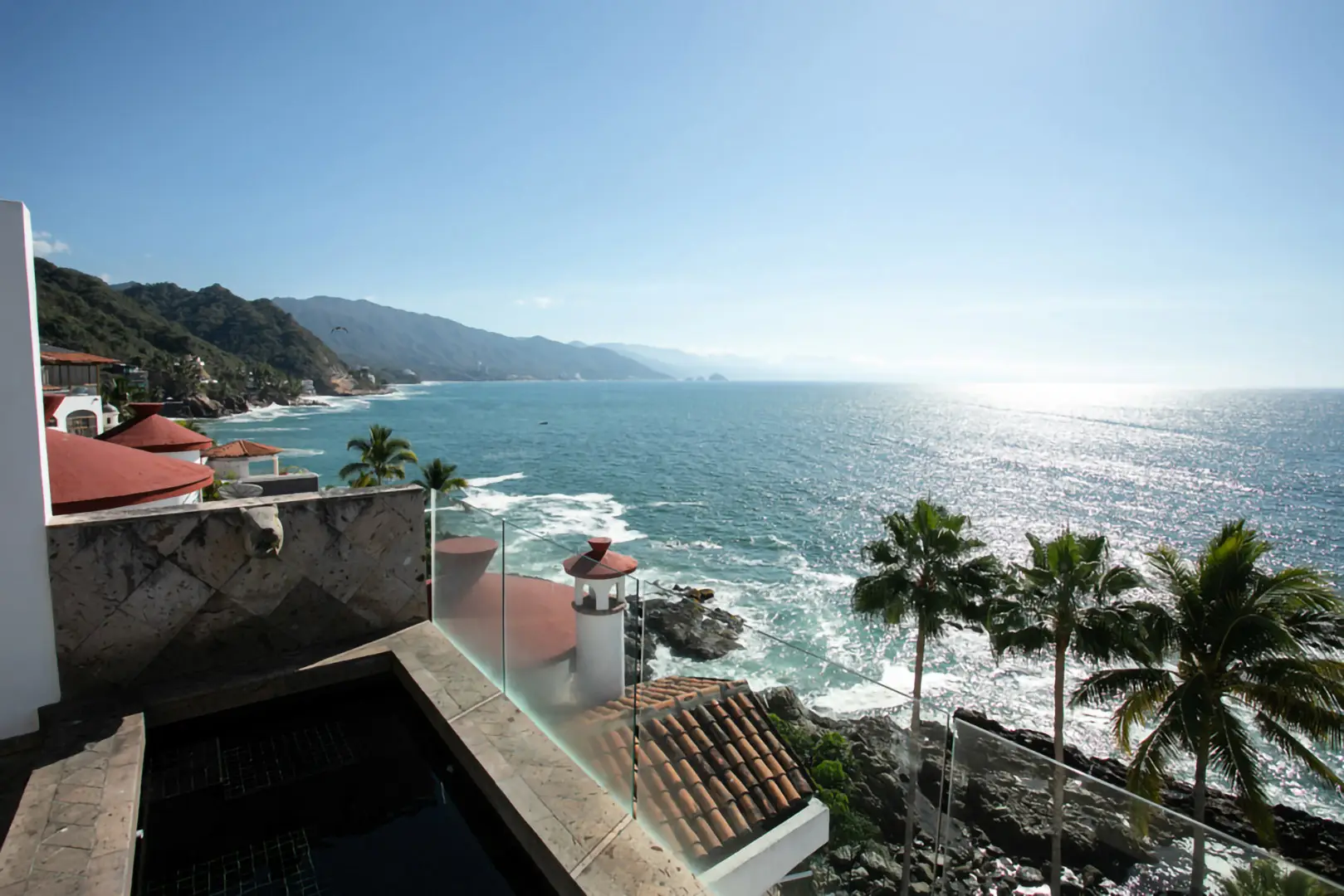 Ocean view from terrace with small pool, palm trees, coastal houses, mountains, and clear blue sky.