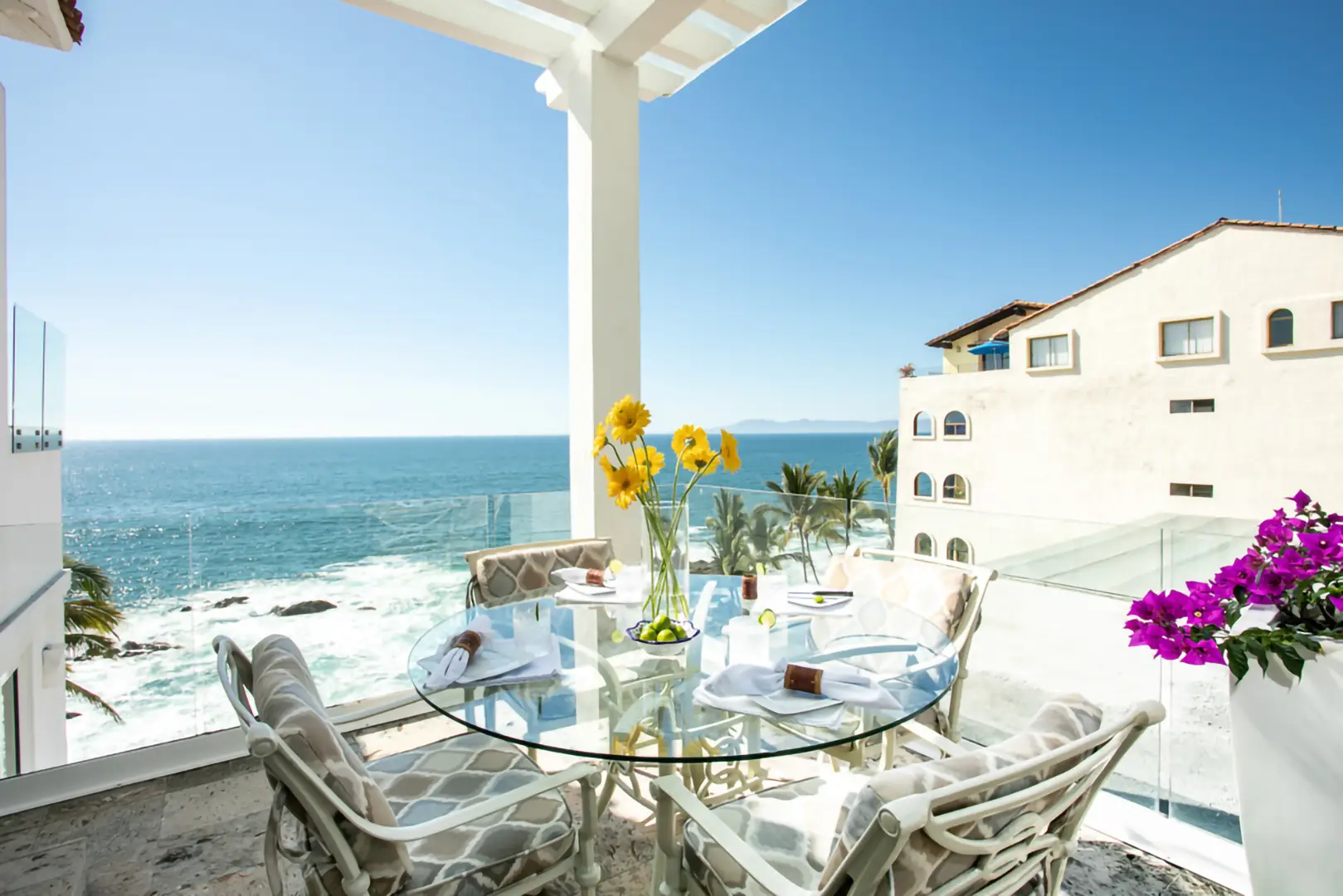 A balcony with a glass table set for four and yellow flowers, overlooking ocean waves, palm trees, and a beige building.
