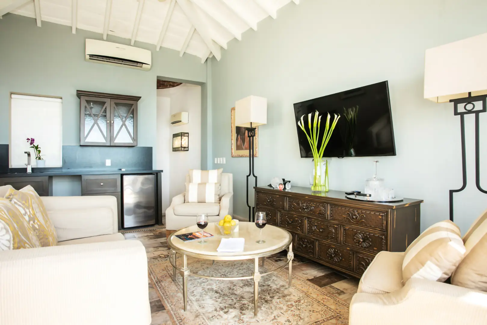 Living room with white sofas, round coffee table with wine glasses and fruit, TV, dresser with flowers, vaulted ceiling.