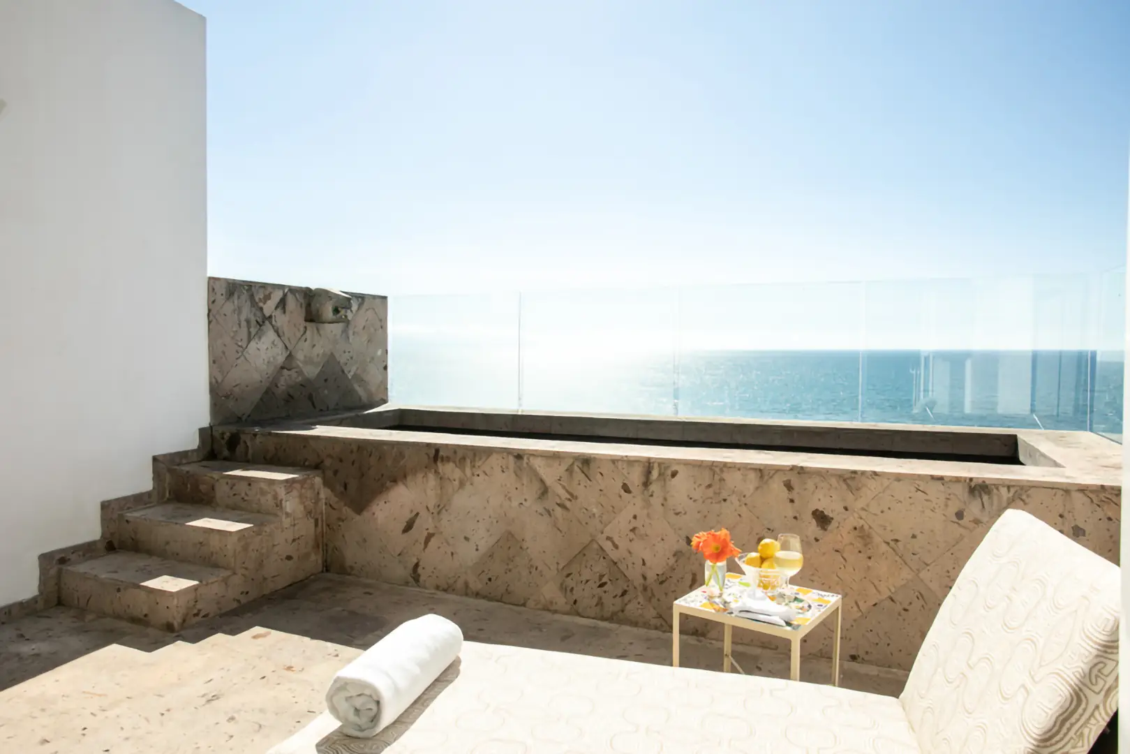 Terrace with small stone plunge pool, glass railing, ocean view, lounge chair, towel, table with flowers and drinks.