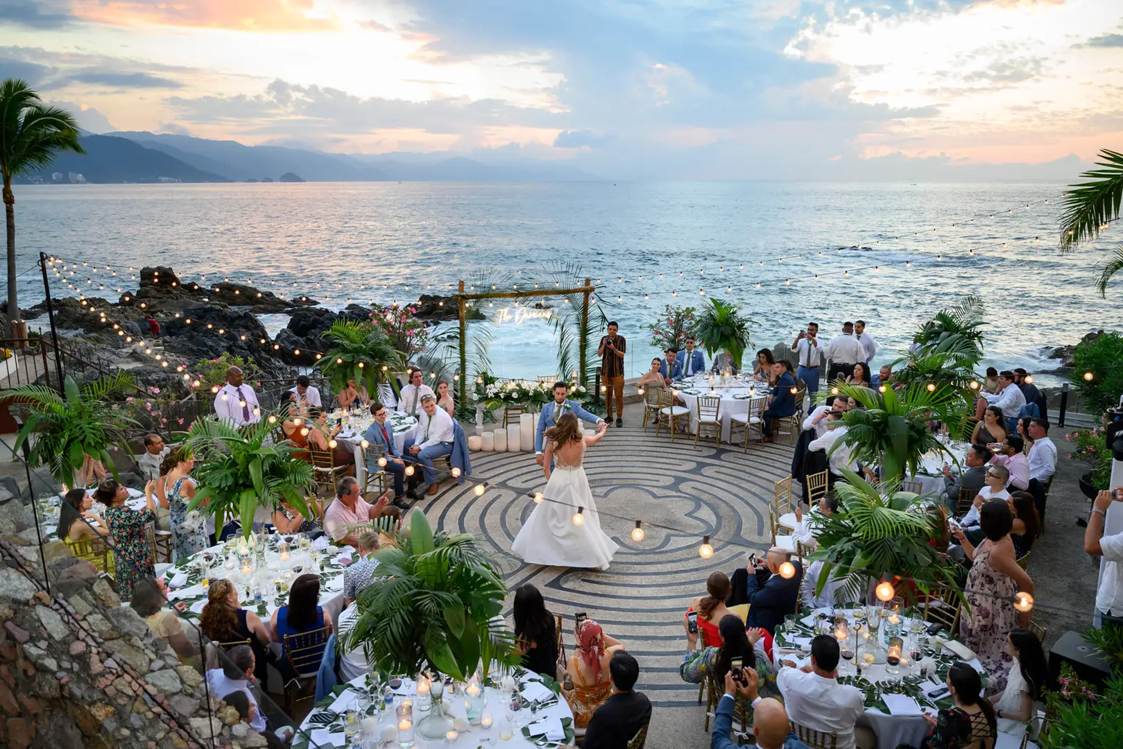 Bride and groom dance on a spiral stone patio, surrounded by guests, tables, tropical plants, lights, ocean and sunset.