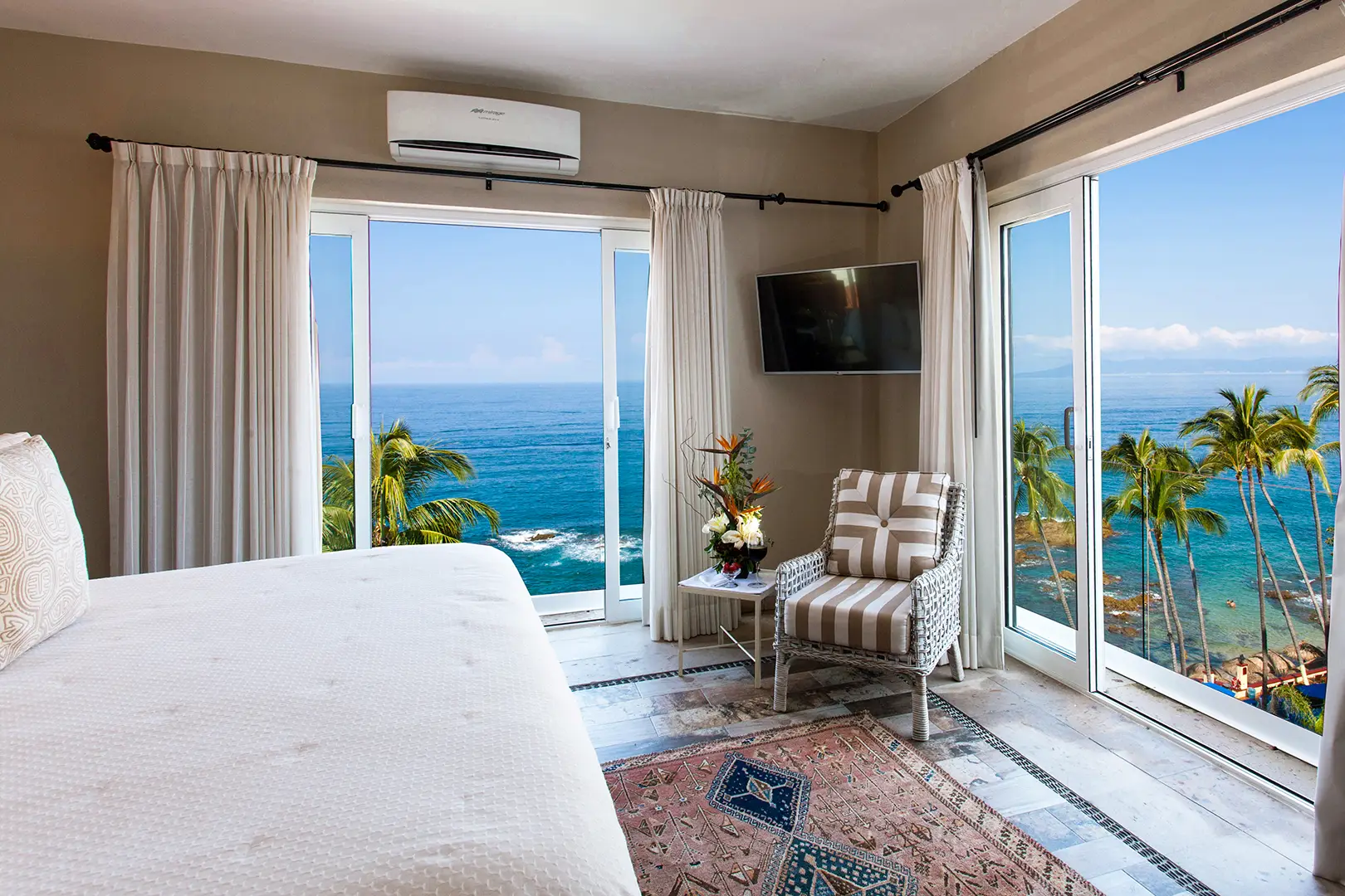 Bedroom with large windows, ocean view, palm trees, bed, patterned chair, side table with flowers, rug, and TV.