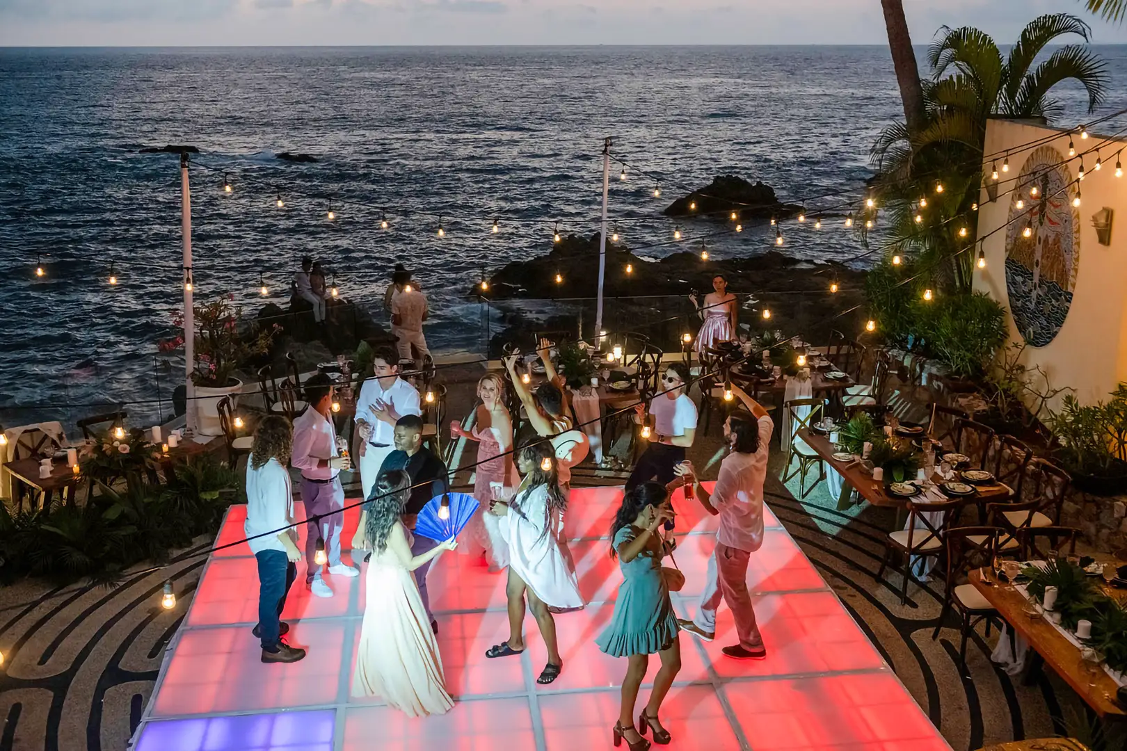 People dancing on a glowing, colorful outdoor dance floor by the ocean at sunset, with string lights and tropical plants.