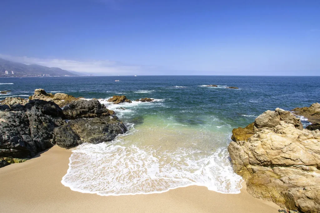 Waves wash onto a sandy beach bordered by rocks, with blue ocean, clear sky, distant mountains and buildings visible.