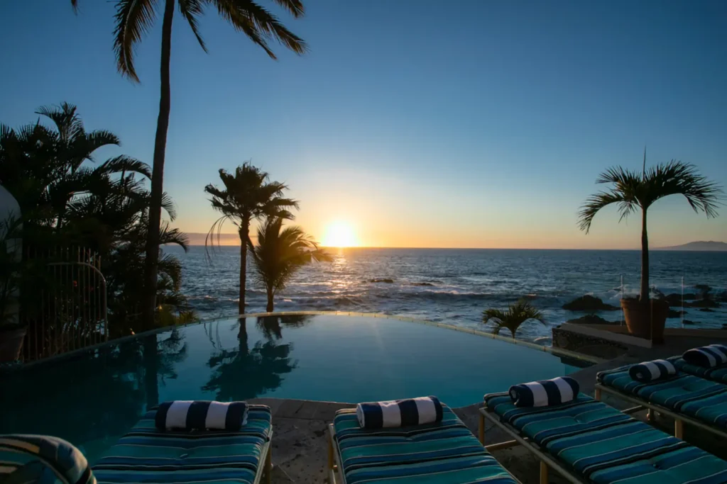 Lounge chairs with striped towels by an infinity pool, ocean view at sunset, palm trees and clear sky visible.
