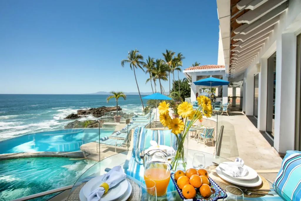Breakfast table with orange juice and fruit overlooking an infinity pool, palm trees, and ocean under blue sky.