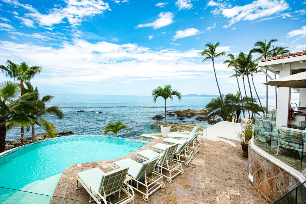 Lounge chairs on a stone patio beside an infinity pool, palm trees, white villa, and ocean under a blue sky with clouds.
