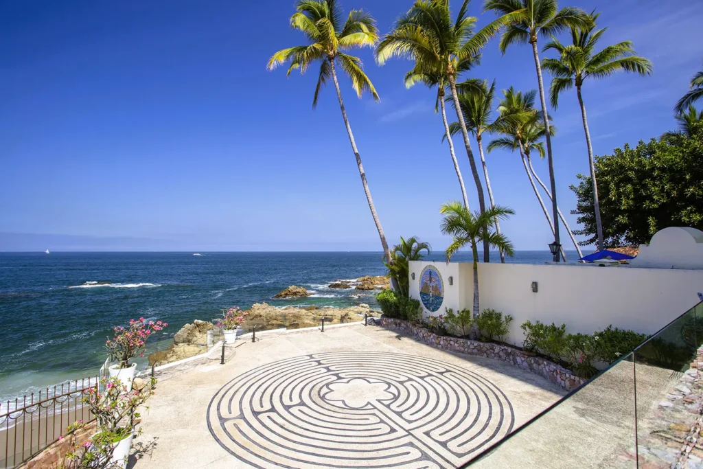 Terrace with circular labyrinth pattern, white wall, palm trees, rocky shore, and blue ocean under clear sky.